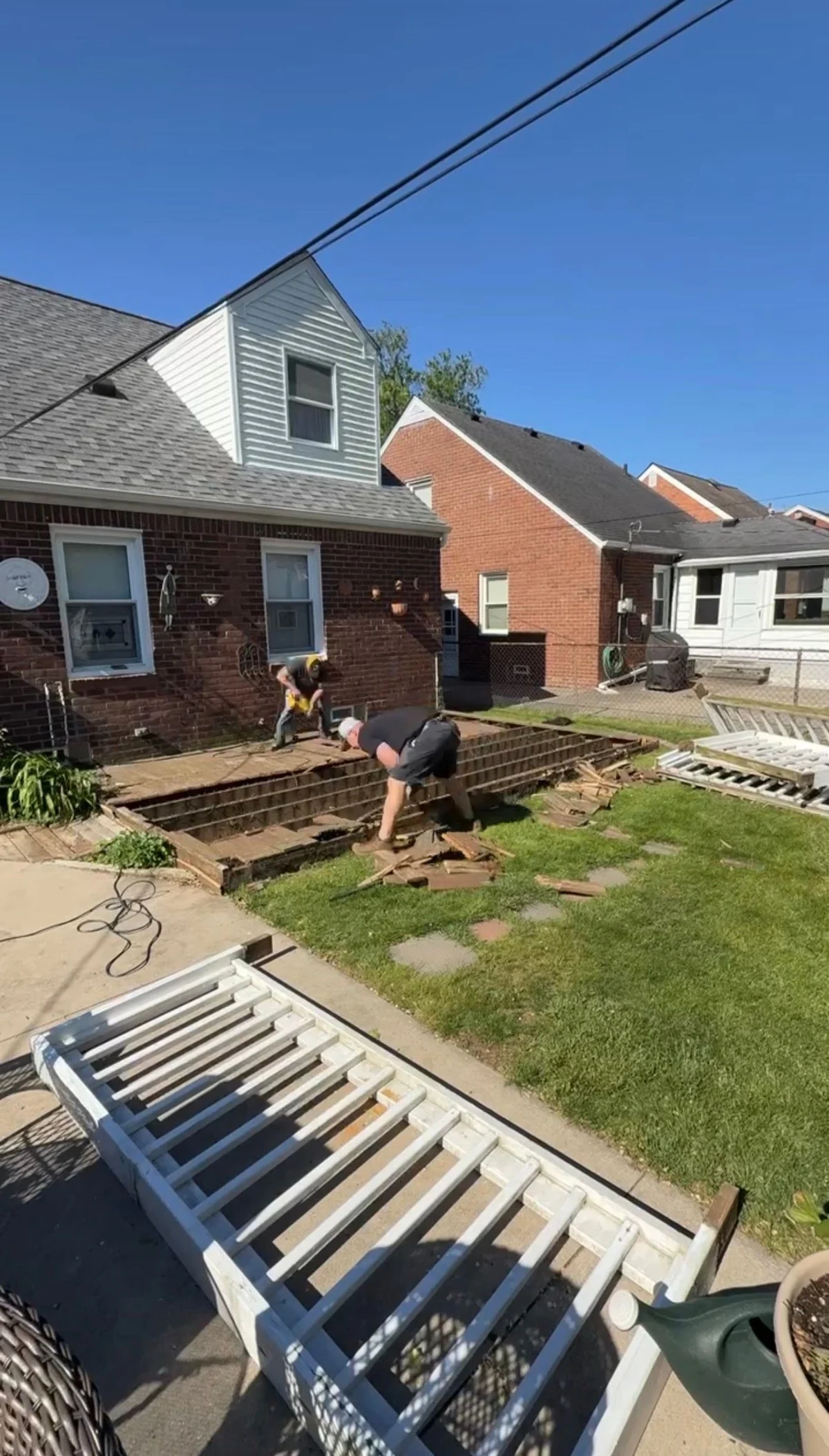 Two workers removing wood deck boards in Grosse Pointe Woods with the underlying framing exposed and removed boards piled on the grass